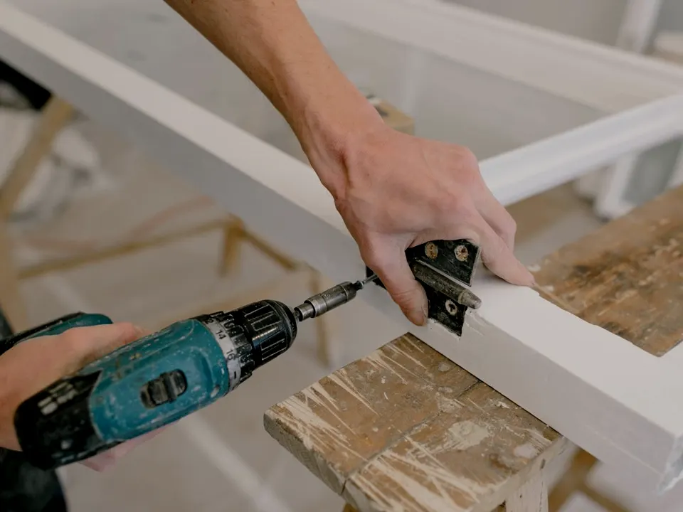 A carpenter screwing a hinge onto a white timber frame with a cordless drill